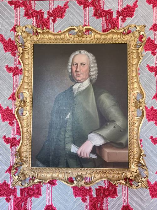 A formal portrait of a man in 18th-century attire, framed in an ornate gold frame against a patterned red wallpaper.