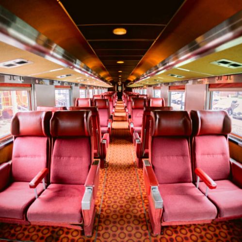 Interior of a vintage train car featuring rows of red upholstered seats and patterned carpet.