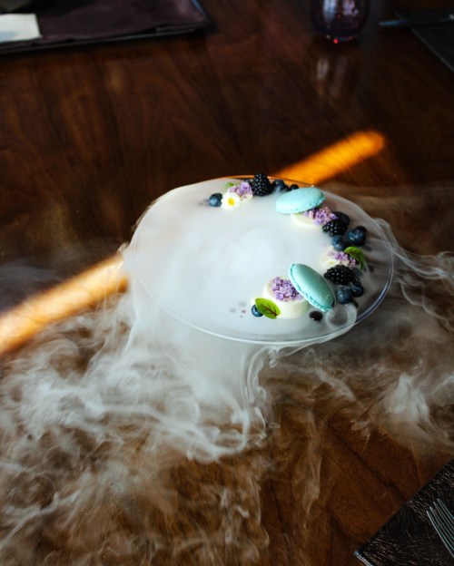 A dessert plate with colorful macarons and berries, surrounded by swirling smoke on a wooden table.
