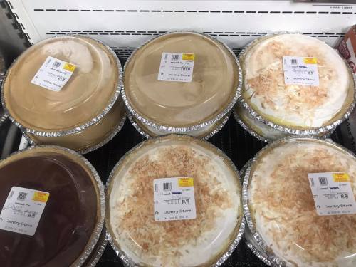 Six pies in aluminum pans, featuring chocolate, coconut, and cream flavors, displayed on a store shelf.