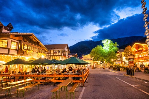 A charming street scene at dusk, featuring illuminated buildings, outdoor seating, and a vibrant tree under a cloudy sky.