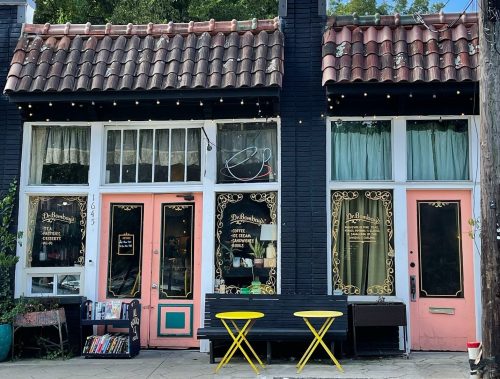 Charming storefront with black and pink doors, large windows, and yellow chairs outside, surrounded by greenery.