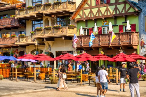 A vibrant street scene with colorful buildings, outdoor dining, and people enjoying the atmosphere under red umbrellas.