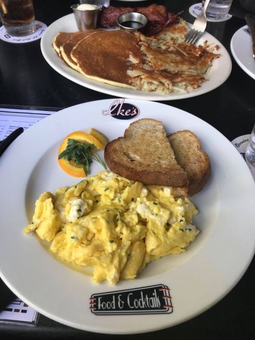 A plate of scrambled eggs with toast, garnished with an orange slice, alongside pancakes and hash browns in the background.