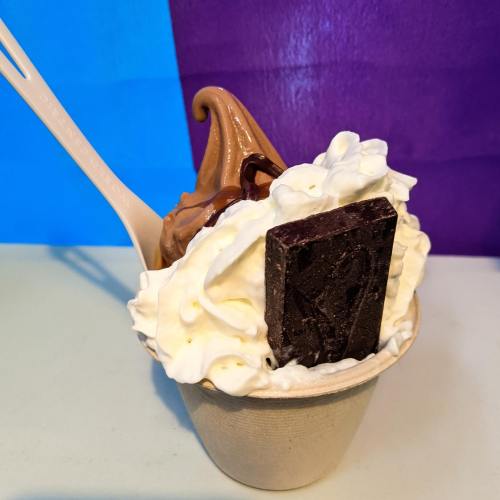A cup of chocolate soft serve topped with whipped cream and a chocolate square, with a wooden spoon beside it.