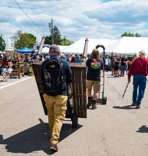 A person pushes a cart loaded with wooden pallets through a busy outdoor market with tents and crowds in the background.