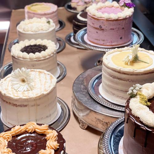 A display of various decorated cakes on silver platters, featuring different flavors and floral accents.