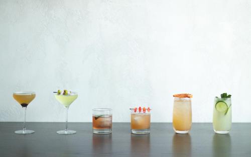 A lineup of six colorful cocktails on a wooden surface against a light wall.