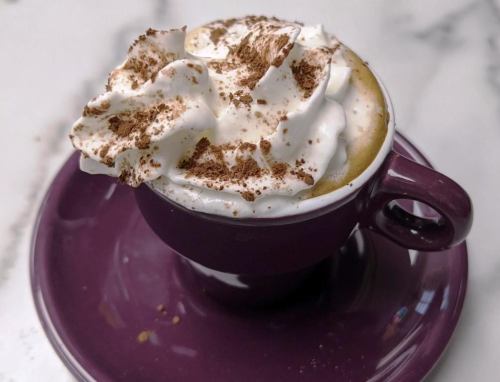 A close-up of a purple cup filled with coffee topped with whipped cream and chocolate shavings, resting on a matching saucer.