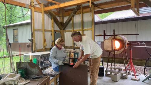 A man and woman work together in a workshop, shaping metal with tools and a furnace in the background.