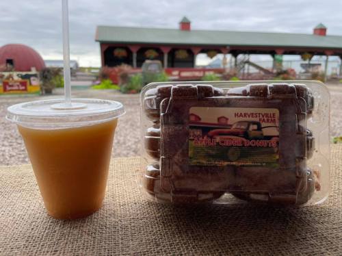 A clear cup of apple cider next to a package of apple cider donuts, with a farm backdrop.
