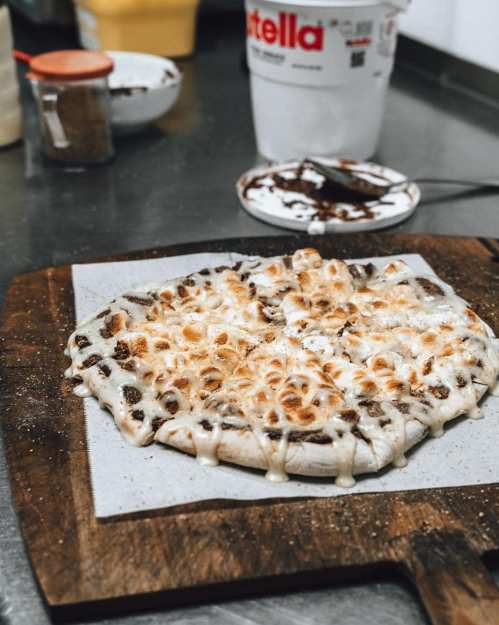 A freshly baked dessert pizza topped with melted marshmallows and drizzled with chocolate on a wooden cutting board.
