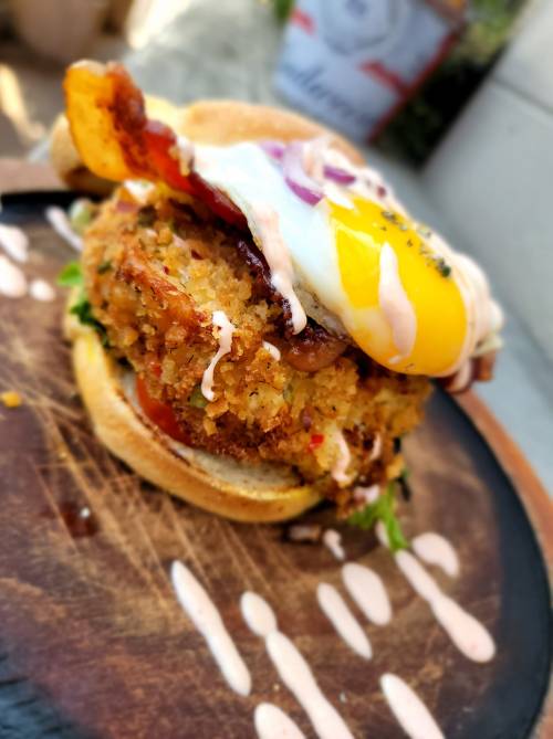 A close-up of a delicious burger with a crispy patty, fried egg, lettuce, and drizzled sauce on a wooden plate.