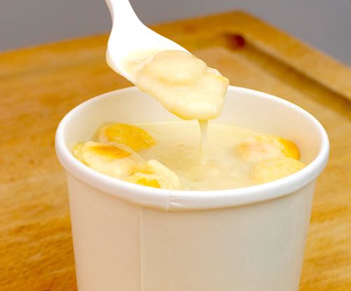 A spoon lifting a creamy dessert with fruit pieces from a white cup, set on a wooden surface.