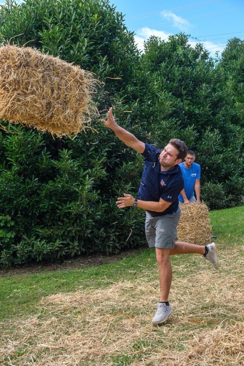 A man throws a hay bale while another man stands behind him, surrounded by greenery and straw on the ground.