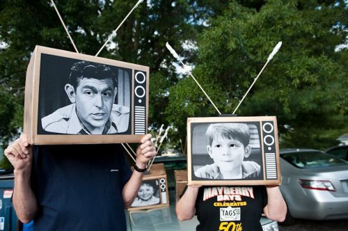Two people with cardboard TV heads featuring black-and-white images of a man and a boy, standing outdoors.