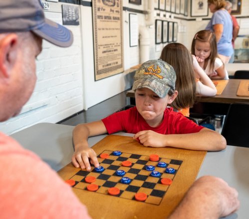 A young boy in a cap concentrates on a checkers game against an adult, with other children playing in the background.