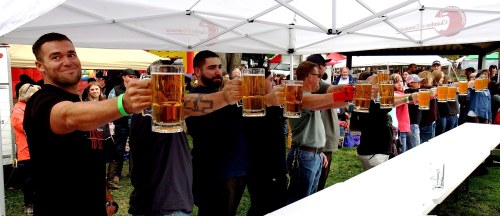 A group of people raising large beer mugs in a festive outdoor setting, with a crowd in the background.