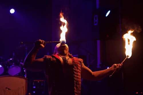 A performer breathes fire during a dramatic stage act, illuminated by colorful lights.