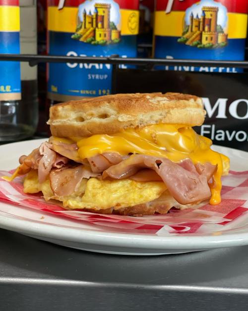A hearty breakfast sandwich with a biscuit, cheese, ham, and an egg, served on a plate with colorful syrup bottles in the background.