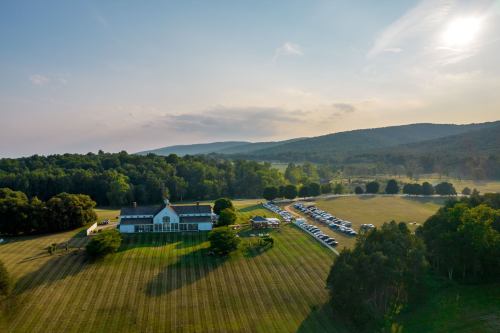 Aerial view of a large green field with a white building and parked cars, surrounded by trees and rolling hills.