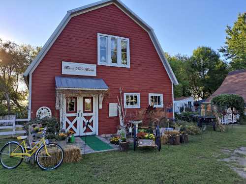 A red barn-style building with a white door, surrounded by flowers and decorative items, set in a green landscape.