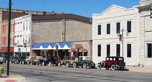 A row of vintage cars parked along a street with historic buildings in the background under a clear blue sky.