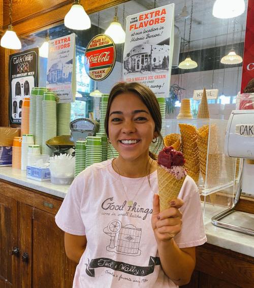 A smiling young woman holds a cone of ice cream in a colorful ice cream shop with various toppings and signs in the background.