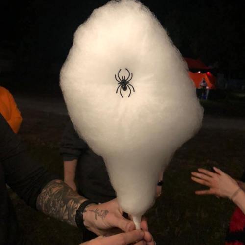 A person holds a cotton candy cone with a black spider decoration on it, set against a dark background.