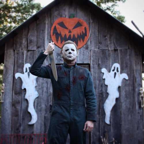 A masked figure holding a knife stands in front of a spooky cabin with a pumpkin and ghost decorations.