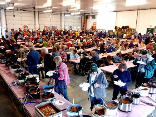A large crowd gathers in a hall for a chili cook-off, with tables filled with various chili dishes and attendees serving themselves.