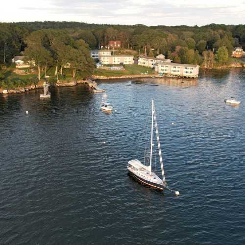 Aerial view of a calm lake with boats and a shoreline featuring green trees and houses in the background.