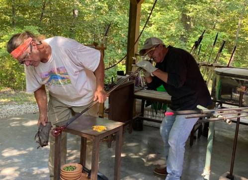 Two glassblowers work outdoors, one shaping glass while the other uses a blowpipe, surrounded by tools and greenery.