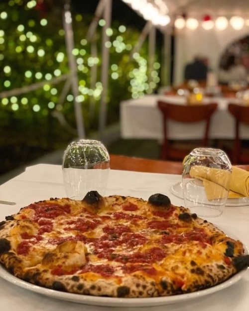 A large pizza with a golden crust and tomato topping sits on a table, surrounded by soft, glowing lights.