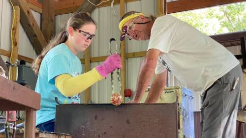 A young girl and an older man work together in a workshop, focusing on a glassblowing project.