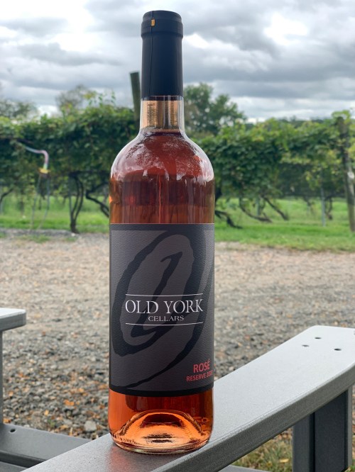A bottle of Old York Cellars rosé wine sits on a table, with vineyards and cloudy skies in the background.
