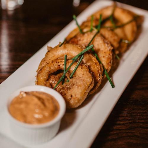 A plate of golden-brown fried onion rings garnished with chives, served with a small bowl of dipping sauce.
