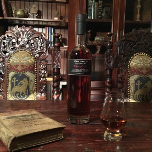 A bottle of whiskey labeled "Abomination" sits on a table next to a glass and an old book, with ornate wooden chairs in the background.