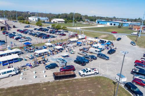 Aerial view of a busy outdoor market with tents, vendors, and parked cars in a large lot on a sunny day.