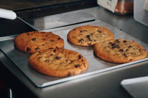 Four freshly baked chocolate chip cookies on a baking tray, with a golden-brown color and a soft texture.