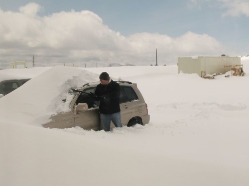 A person stands next to a car partially buried in deep snow, with snowdrifts surrounding the vehicle under a cloudy sky.