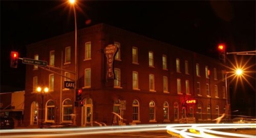 A brick hotel at night, illuminated by streetlights, with a café sign visible and traffic lights glowing red.