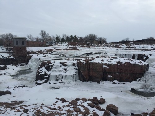 A frozen waterfall with icy rocks and a cloudy sky in the background, surrounded by trees and a building.