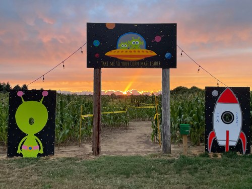 A colorful corn maze entrance featuring alien and rocket artwork, with a sunset in the background.