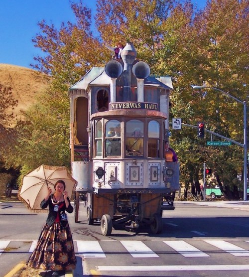A woman in a vintage dress holds an umbrella in front of a whimsical, steampunk-style vehicle on a sunny street.