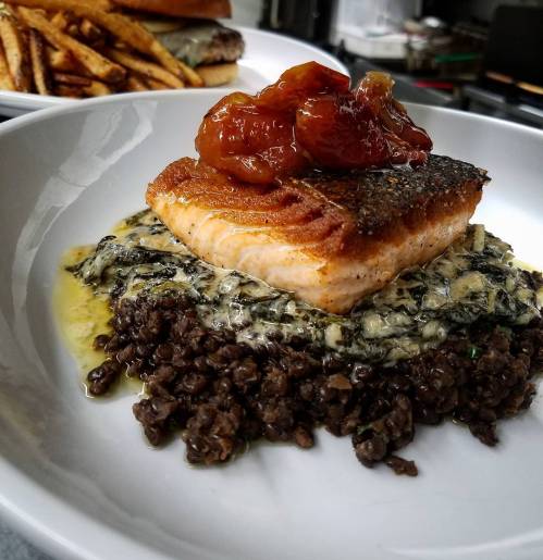 A plate featuring seared salmon on a bed of creamy lentils, topped with a savory sauce and accompanied by fries in the background.