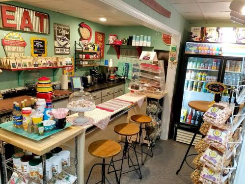 Cozy café interior with a counter, stools, snacks, and a display fridge filled with drinks and treats.