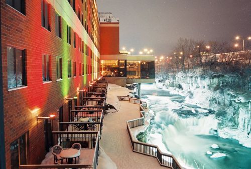 A snowy riverside view at night, featuring a hotel with colorful lights and a flowing waterfall nearby.