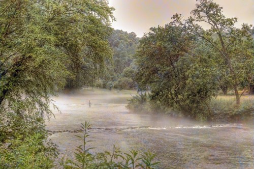 A misty river scene surrounded by lush trees, with a lone figure standing in the water.