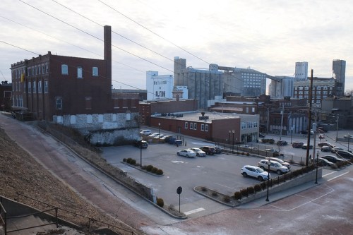 A view of an industrial area featuring brick buildings, a parking lot, and silos in the background under a cloudy sky.